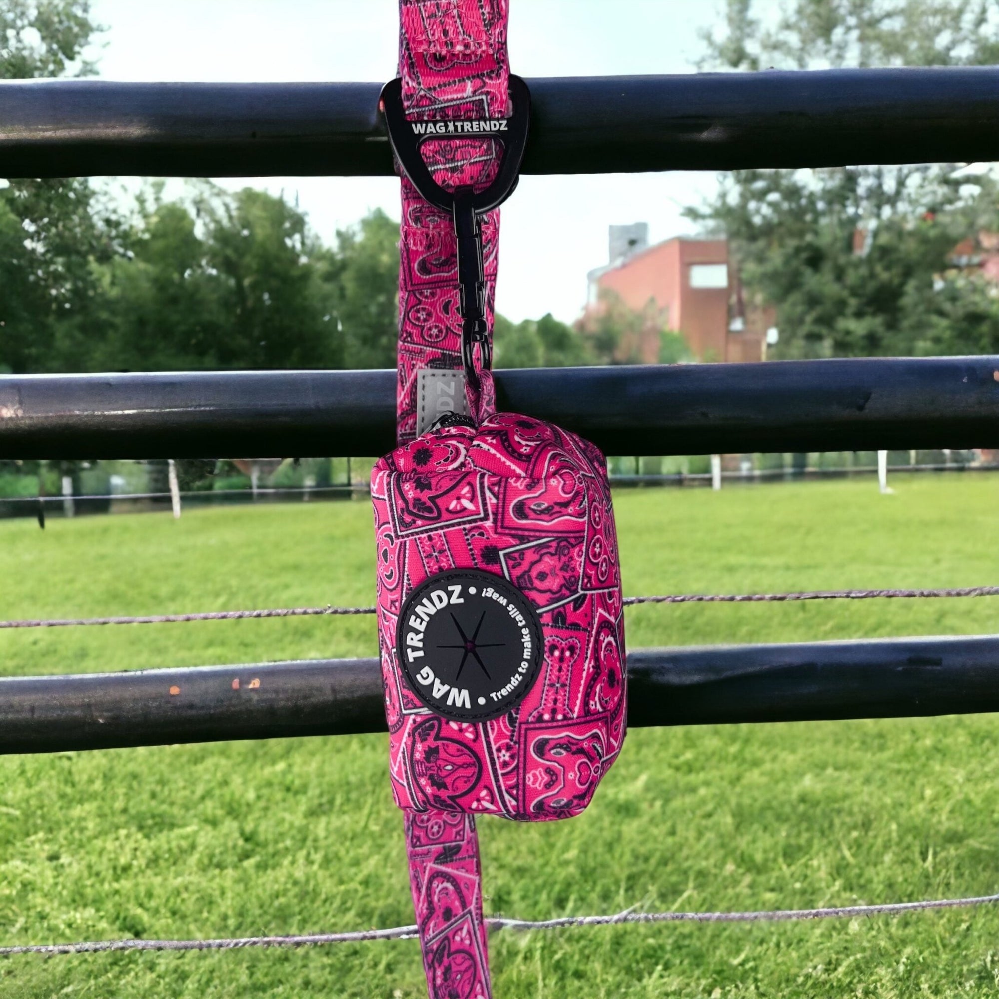 Dog Poo Bag Holder - Bandana Boujee - Hot Pink with black zipper and black rubber logo dispenser on front hanging on a matching leash outdoors on a black fence with green grass in background - Wag Trendz