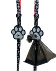 Poop Buddy - black and gray resin dog paw - two hanging on two black & white dog leashes XO with red accents and black plastic bag carrying poop pushed through hole on one - against white background - Wag Trendz
